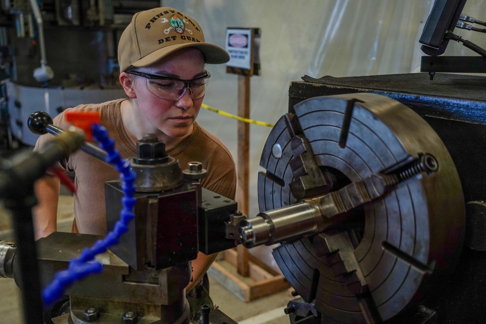 A young person wearing safety glasses and a tan baseball cap is operating a metal lathe in what appears to be a machine shop or workshop. They are closely inspecting the work piece mounted in a large four-jaw chuck on the lathe. The image captures focused, hands-on machining work in an industrial setting.