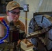 A young person wearing safety glasses and a tan baseball cap is operating a metal lathe in what appears to be a machine shop or workshop. They are closely inspecting the work piece mounted in a large four-jaw chuck on the lathe. The image captures focused, hands-on machining work in an industrial setting.