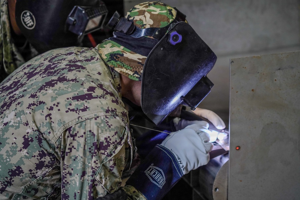 A military personnel wearing digital camouflage uniform and a camouflage cap is performing welding work on a metal surface. The individual is equipped with a welding helmet/face shield and heavy-duty protective gloves, actively producing a visible weld arc. A second person in military uniform is partially visible in the background observing the work.