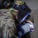 A military personnel wearing digital camouflage uniform and a camouflage cap is performing welding work on a metal surface. The individual is equipped with a welding helmet/face shield and heavy-duty protective gloves, actively producing a visible weld arc. A second person in military uniform is partially visible in the background observing the work.