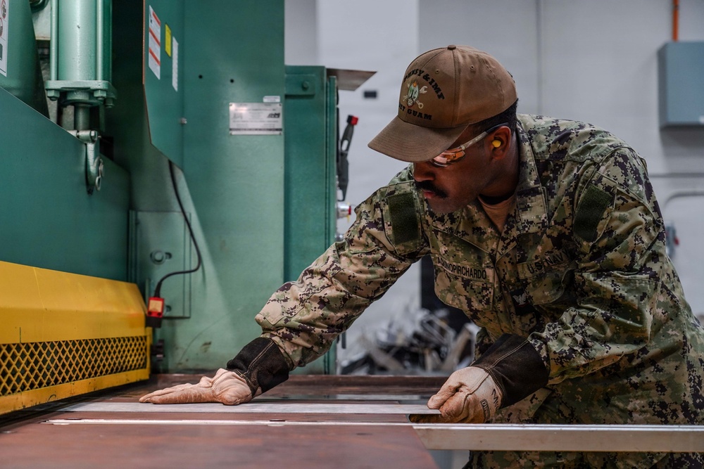 A U.S. Navy sailor in NWU Type III camouflage uniform is working with sheet metal at an industrial machine, likely a press brake or metal shear. The sailor is wearing protective gloves and ear protection while carefully positioning a metal sheet against the machine. The setting appears to be a military fabrication or machine shop facility.