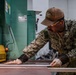 A U.S. Navy sailor in NWU Type III camouflage uniform is working with sheet metal at an industrial machine, likely a press brake or metal shear. The sailor is wearing protective gloves and ear protection while carefully positioning a metal sheet against the machine. The setting appears to be a military fabrication or machine shop facility.
