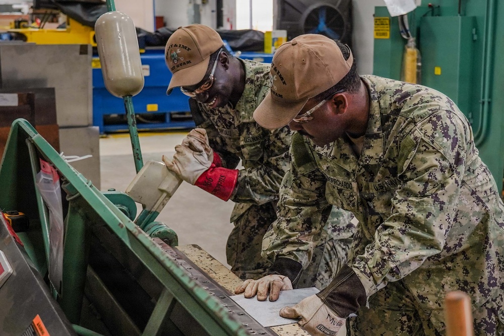 Two U.S. Navy personnel in NWU Type III (AOR-2) camouflage uniforms are working together in what appears to be a military maintenance or fabrication shop. Both are wearing tan caps with 'IMF GUAM' text and protective gloves while operating or inspecting a green metal bending/brake press machine. The name tape 'PIRICHARDO' is visible on the right individual's uniform.