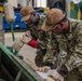 Two U.S. Navy personnel in NWU Type III (AOR-2) camouflage uniforms are working together in what appears to be a military maintenance or fabrication shop. Both are wearing tan caps with 'IMF GUAM' text and protective gloves while operating or inspecting a green metal bending/brake press machine. The name tape 'PIRICHARDO' is visible on the right individual's uniform.