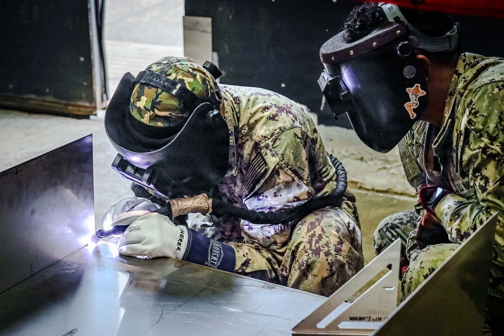 Two military personnel in camouflage uniforms are performing welding work on metal components. The individual on the left is actively welding, producing a visible arc flash, while the person on the right observes closely. Both are wearing protective welding helmets and the welder is wearing heavy-duty protective gloves.