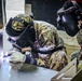Two military personnel in camouflage uniforms are performing welding work on metal components. The individual on the left is actively welding, producing a visible arc flash, while the person on the right observes closely. Both are wearing protective welding helmets and the welder is wearing heavy-duty protective gloves.