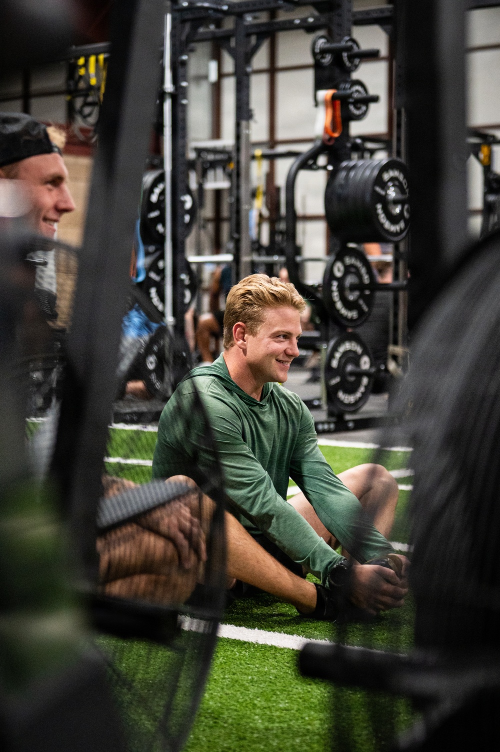 A young man with blonde hair wearing a green long-sleeve hoodie is seated on artificial turf flooring inside a weight training facility, smiling and appearing relaxed. The gym is well-equipped with multiple power racks loaded with barbells and weight plates visible in the background. The photo is taken from a low angle through gym equipment, giving a candid, immersive perspective.
