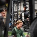 A young man with blonde hair wearing a green long-sleeve hoodie is seated on artificial turf flooring inside a weight training facility, smiling and appearing relaxed. The gym is well-equipped with multiple power racks loaded with barbells and weight plates visible in the background. The photo is taken from a low angle through gym equipment, giving a candid, immersive perspective.