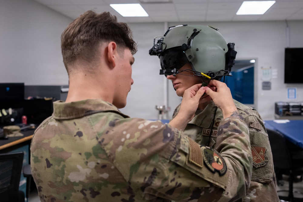 Two U.S. Air Force personnel in OCP (Operational Camouflage Pattern) uniforms are shown in what appears to be an indoor training or equipment facility. One service member is assisting the other with fitting or adjusting a military flight/tactical helmet equipped with night vision or display mounts. The setting appears to be a technical or equipment room with computers and monitors visible in the background.