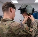 Two U.S. Air Force personnel in OCP (Operational Camouflage Pattern) uniforms are shown in what appears to be an indoor training or equipment facility. One service member is assisting the other with fitting or adjusting a military flight/tactical helmet equipped with night vision or display mounts. The setting appears to be a technical or equipment room with computers and monitors visible in the background.