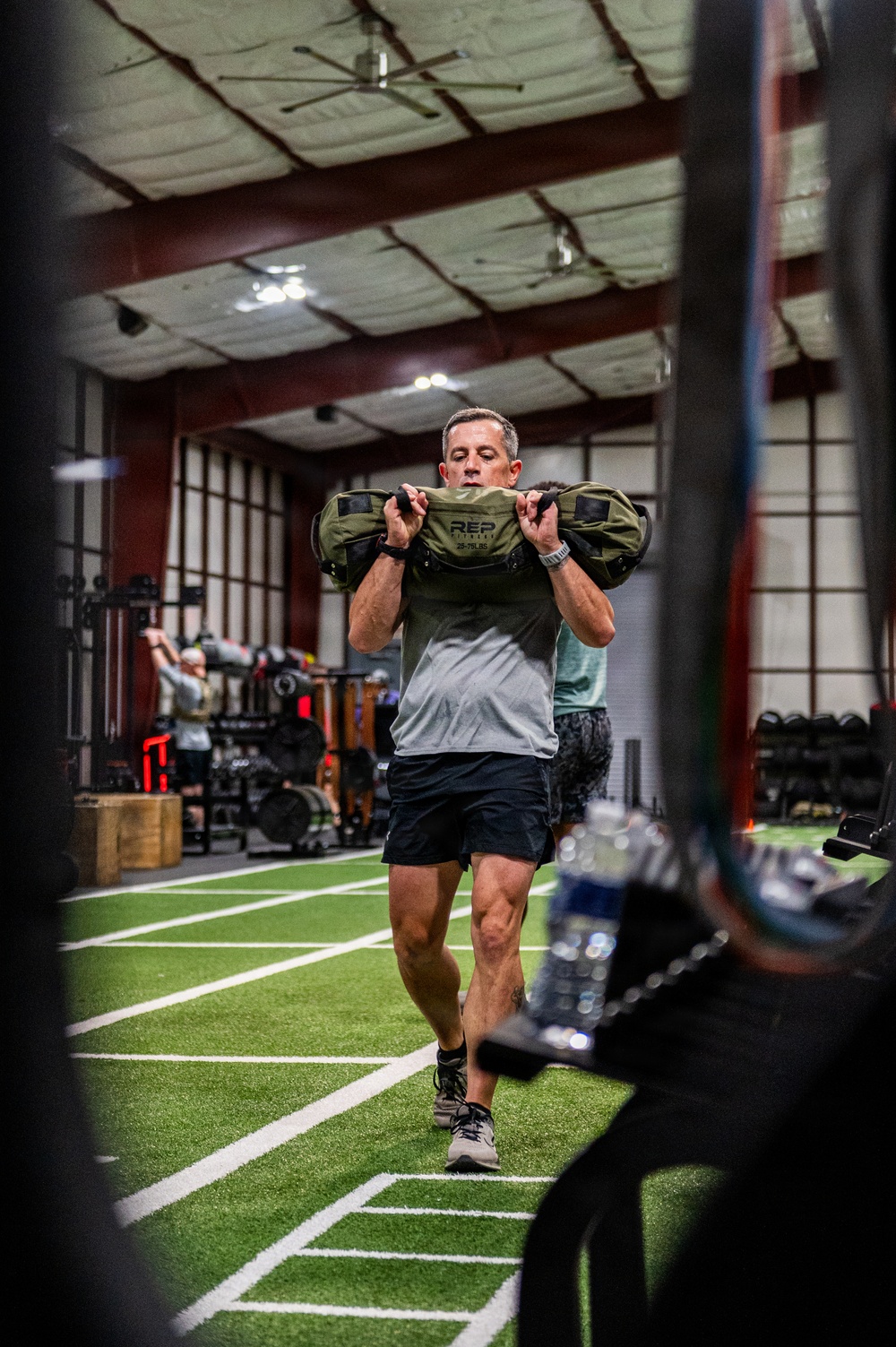 A middle-aged man is performing a sandbag carry exercise on an indoor artificial turf training facility. He is holding an olive-green 'REP Fitness' sandbag at chest level while walking forward. The gym features multiple athletes training simultaneously in a large, well-lit performance facility.