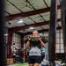 A middle-aged man is performing a sandbag carry exercise on an indoor artificial turf training facility. He is holding an olive-green 'REP Fitness' sandbag at chest level while walking forward. The gym features multiple athletes training simultaneously in a large, well-lit performance facility.