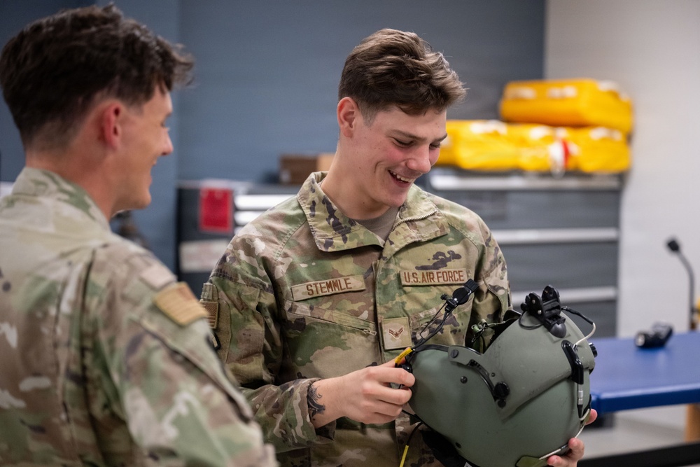 Two U.S. Air Force personnel in OCP (Operational Camouflage Pattern) uniforms are interacting in what appears to be an equipment maintenance or supply room. The airman facing the camera, identified by name tape as 'STEMMLE,' is smiling while handling a military flight helmet equipped with attachments. The setting suggests a technical or life support equipment workspace.