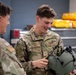 Two U.S. Air Force personnel in OCP (Operational Camouflage Pattern) uniforms are interacting in what appears to be an equipment maintenance or supply room. The airman facing the camera, identified by name tape as 'STEMMLE,' is smiling while handling a military flight helmet equipped with attachments. The setting suggests a technical or life support equipment workspace.