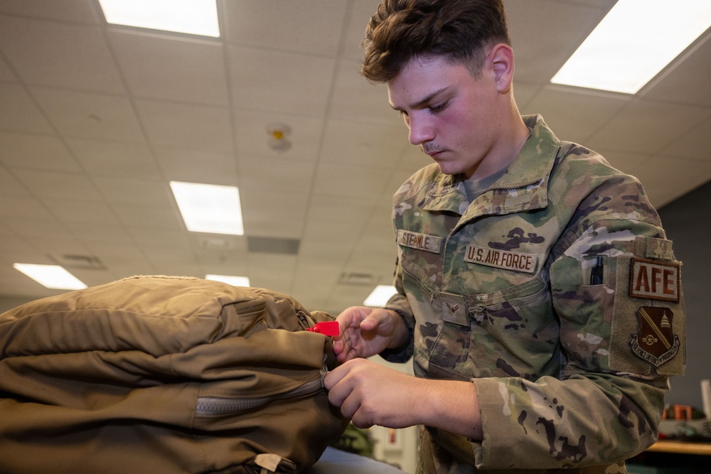 A U.S. Air Force airman wearing OCP (Operational Camouflage Pattern) uniform is inspecting or handling military gear, specifically working with a zipper on what appears to be a survival vest or equipment bag. The name tape reads 'STERMLE' and the right sleeve displays an 'AFE' (Aircrew Flight Equipment) patch, indicating a specialty role. The setting appears to be an indoor military facility or equipment room.