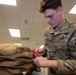 A U.S. Air Force airman wearing OCP (Operational Camouflage Pattern) uniform is inspecting or handling military gear, specifically working with a zipper on what appears to be a survival vest or equipment bag. The name tape reads 'STERMLE' and the right sleeve displays an 'AFE' (Aircrew Flight Equipment) patch, indicating a specialty role. The setting appears to be an indoor military facility or equipment room.