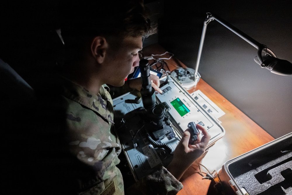 A soldier in MultiCam/OCP camouflage uniform is working with military electronic equipment at a dimly lit desk illuminated by a single desk lamp. The soldier appears to be operating or inspecting a piece of military communications or optical equipment labeled 'ANV-126A' or similar designation. The image is taken from an elevated angle in a low-light environment.