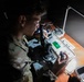A soldier in MultiCam/OCP camouflage uniform is working with military electronic equipment at a dimly lit desk illuminated by a single desk lamp. The soldier appears to be operating or inspecting a piece of military communications or optical equipment labeled 'ANV-126A' or similar designation. The image is taken from an elevated angle in a low-light environment.