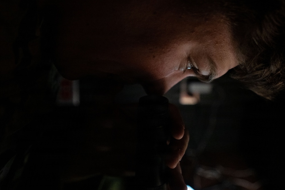 A close-up, low-light photograph of a person's face partially illuminated, appearing to look through or hold a dark optical or mechanical device. The image is heavily shadowed with only portions of the face — particularly around the eye and forehead — catching ambient light. The overall composition is dramatic and moody, with minimal environmental context visible.