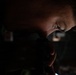 A close-up, low-light photograph of a person's face partially illuminated, appearing to look through or hold a dark optical or mechanical device. The image is heavily shadowed with only portions of the face — particularly around the eye and forehead — catching ambient light. The overall composition is dramatic and moody, with minimal environmental context visible.