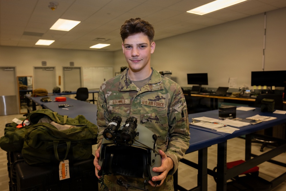 A U.S. Air Force service member named Stemmle is posing in what appears to be a training or equipment room. He is holding night vision goggle (NVG) equipment and wearing OCP (Operational Camouflage Pattern) uniform. The setting appears to be an indoor military facility with workbenches and various equipment visible in the background.