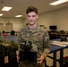 A U.S. Air Force service member named Stemmle is posing in what appears to be a training or equipment room. He is holding night vision goggle (NVG) equipment and wearing OCP (Operational Camouflage Pattern) uniform. The setting appears to be an indoor military facility with workbenches and various equipment visible in the background.