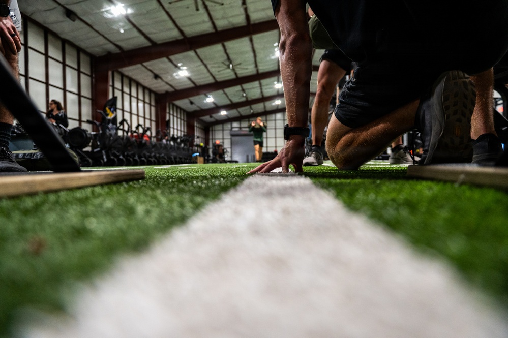 A ground-level photograph taken on an indoor artificial turf facility, showing a person in a crouched/starting position with their hand touching a white yard line. The low-angle perspective emphasizes the turf surface and white stripe running toward the background. The facility appears to be an indoor sports training center equipped with exercise bikes and other fitness equipment.
