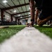 A ground-level photograph taken on an indoor artificial turf facility, showing a person in a crouched/starting position with their hand touching a white yard line. The low-angle perspective emphasizes the turf surface and white stripe running toward the background. The facility appears to be an indoor sports training center equipped with exercise bikes and other fitness equipment.
