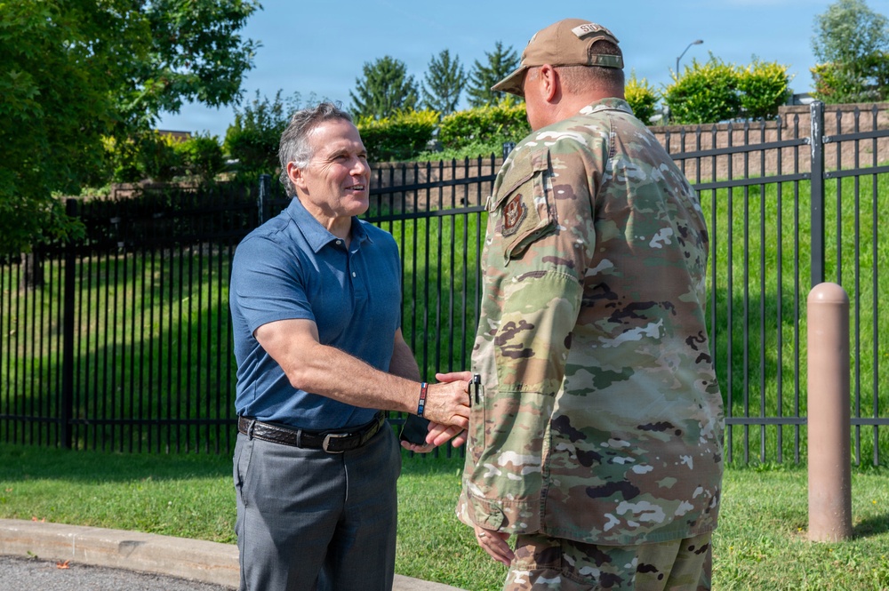 A civilian man in a blue polo shirt is shaking hands with a U.S. military service member in OCP (Operational Camouflage Pattern) uniform outdoors. The setting appears to be a military installation, evidenced by the security fencing and well-maintained grounds. The interaction appears cordial and formal, suggesting an official greeting or visit.