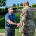 A civilian man in a blue polo shirt is shaking hands with a U.S. military service member in OCP (Operational Camouflage Pattern) uniform outdoors. The setting appears to be a military installation, evidenced by the security fencing and well-maintained grounds. The interaction appears cordial and formal, suggesting an official greeting or visit.