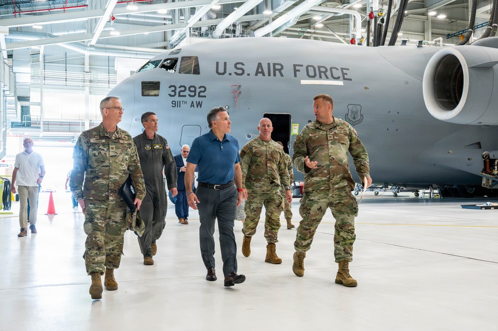 A group of U.S. Air Force military personnel in OCP (Operational Camouflage Pattern) uniforms and one civilian in civilian attire are walking through a military aircraft hangar. Behind them is a large U.S. Air Force transport aircraft, identified as tail number 3292 belonging to the 911th Airlift Wing. The scene appears to be an official tour or inspection visit.