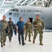 A group of U.S. Air Force military personnel in OCP (Operational Camouflage Pattern) uniforms and one civilian in civilian attire are walking through a military aircraft hangar. Behind them is a large U.S. Air Force transport aircraft, identified as tail number 3292 belonging to the 911th Airlift Wing. The scene appears to be an official tour or inspection visit.