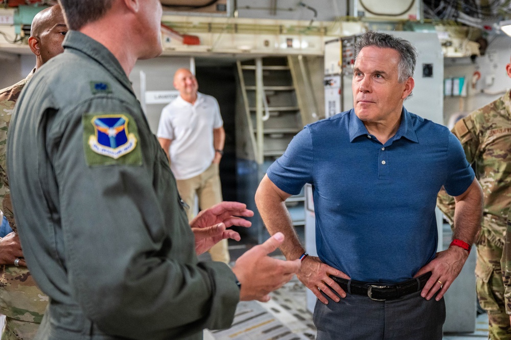 A civilian man in a blue polo shirt is engaged in conversation with a U.S. Air Force officer in an olive green flight suit inside what appears to be a military aircraft or hangar facility. Additional military personnel in camouflage uniforms and a civilian in a white shirt are visible in the background. The setting suggests an official visit or briefing of a civilian dignitary or official to a military installation.