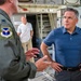 A civilian man in a blue polo shirt is engaged in conversation with a U.S. Air Force officer in an olive green flight suit inside what appears to be a military aircraft or hangar facility. Additional military personnel in camouflage uniforms and a civilian in a white shirt are visible in the background. The setting suggests an official visit or briefing of a civilian dignitary or official to a military installation.