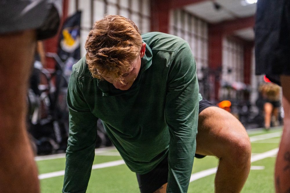 A young male athlete with reddish-blonde hair is bent forward in a fatigued or resting posture, wearing a green long-sleeve shirt and black shorts. The setting appears to be an indoor athletic training facility with artificial turf flooring and red walls. Other individuals are partially visible in the foreground and background.
