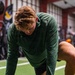 A young male athlete with reddish-blonde hair is bent forward in a fatigued or resting posture, wearing a green long-sleeve shirt and black shorts. The setting appears to be an indoor athletic training facility with artificial turf flooring and red walls. Other individuals are partially visible in the foreground and background.