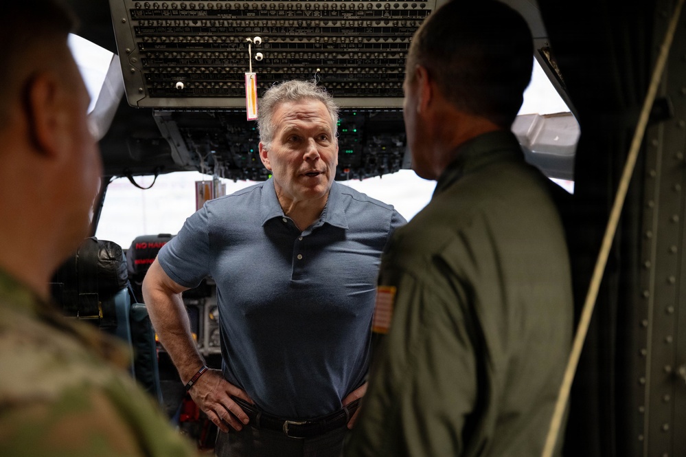 A civilian man in a blue polo shirt stands inside what appears to be a large military aircraft, likely a cargo or transport plane, conversing with two uniformed military personnel. The setting appears to be the interior forward section of the aircraft near the flight deck area. The interaction suggests a briefing, tour, or official visit.