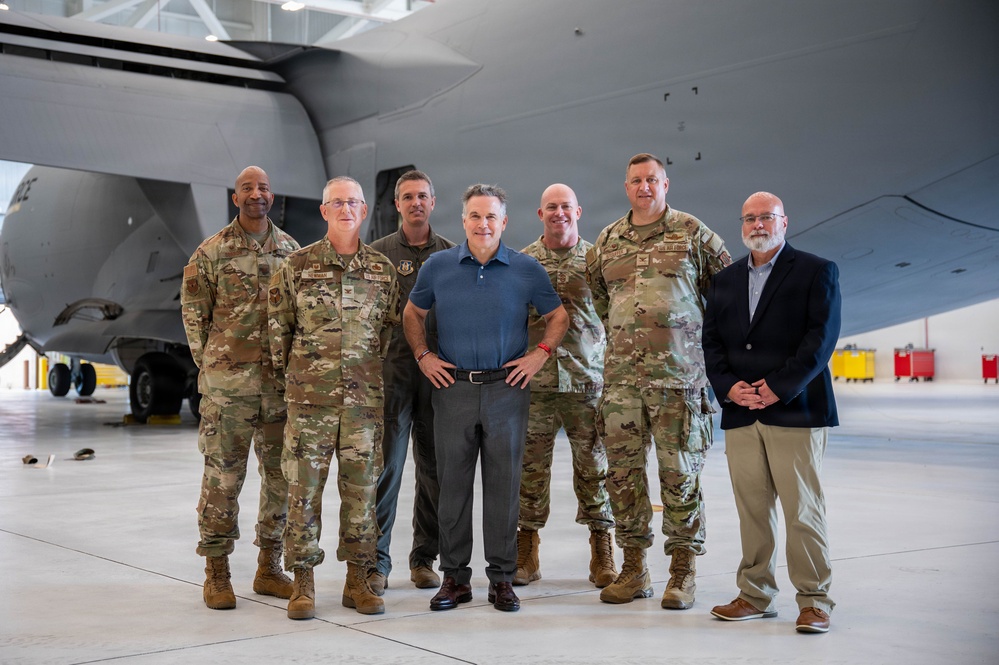 A group of seven individuals pose for a formal group photo inside a military aircraft hangar. Five of the individuals are wearing U.S. Air Force OCP (Operational Camouflage Pattern) uniforms, while two are dressed in civilian attire. They are standing in front of a large gray military aircraft.