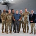 A group of seven individuals pose for a formal group photo inside a military aircraft hangar. Five of the individuals are wearing U.S. Air Force OCP (Operational Camouflage Pattern) uniforms, while two are dressed in civilian attire. They are standing in front of a large gray military aircraft.