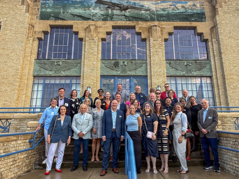 A large group of approximately 25-30 adults poses for a formal group photograph on the steps of an Art Deco-style building. The building features distinctive architectural elements including decorative eagle reliefs, large multi-pane windows, and a mural or relief panel depicting an aircraft near the roofline. The group appears to be attending a professional or civic event, as many individuals are wearing name badges.