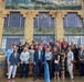 A large group of approximately 25-30 adults poses for a formal group photograph on the steps of an Art Deco-style building. The building features distinctive architectural elements including decorative eagle reliefs, large multi-pane windows, and a mural or relief panel depicting an aircraft near the roofline. The group appears to be attending a professional or civic event, as many individuals are wearing name badges.