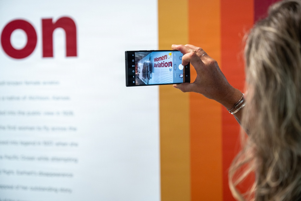 A woman with blonde wavy hair is photographing an exhibition display using a smartphone. The phone screen shows a 'Women in Aviation' exhibit sign, suggesting this is a museum or educational display. The background features a white informational panel with red text and a colorful striped wall in orange and red tones.