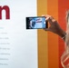 A woman with blonde wavy hair is photographing an exhibition display using a smartphone. The phone screen shows a 'Women in Aviation' exhibit sign, suggesting this is a museum or educational display. The background features a white informational panel with red text and a colorful striped wall in orange and red tones.