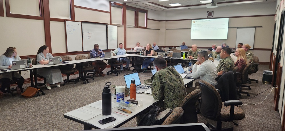 A formal meeting or briefing is taking place in a conference room with a mix of military personnel in uniform and civilians seated around arranged tables in a U-shape configuration. A presentation is being displayed on a projector screen at the front of the room. The setting appears to be a military installation or government facility based on the participants and environment.