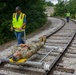 A training or demonstration exercise is taking place on an active railroad track, where a person in military camouflage uniform is lying on a wheeled rail stretcher/litter device. A worker in a high-visibility safety vest is pushing the rail-mounted stretcher along the tracks. Two additional workers in safety vests are visible walking along the tracks in the background.