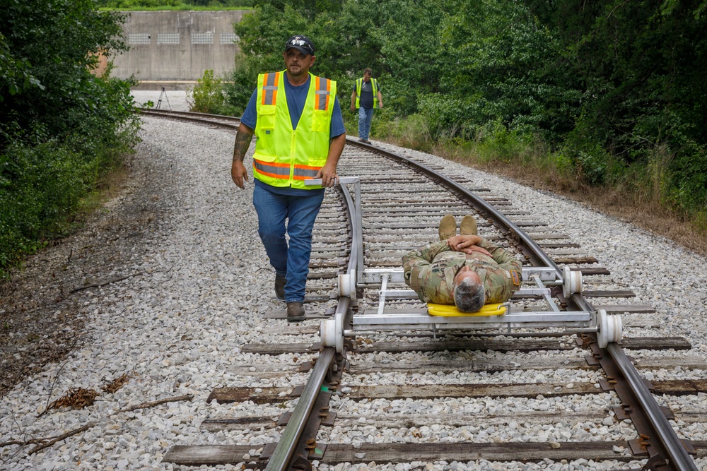A person in military camouflage uniform is lying on a small rail cart (likely a track inspection or maintenance trolley) on a curved railroad track. A worker wearing a high-visibility safety vest and blue jeans walks alongside the cart on the gravel trackbed. A second person in a safety vest is visible further down the track in the background.