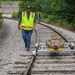 A person in military camouflage uniform is lying on a small rail cart (likely a track inspection or maintenance trolley) on a curved railroad track. A worker wearing a high-visibility safety vest and blue jeans walks alongside the cart on the gravel trackbed. A second person in a safety vest is visible further down the track in the background.