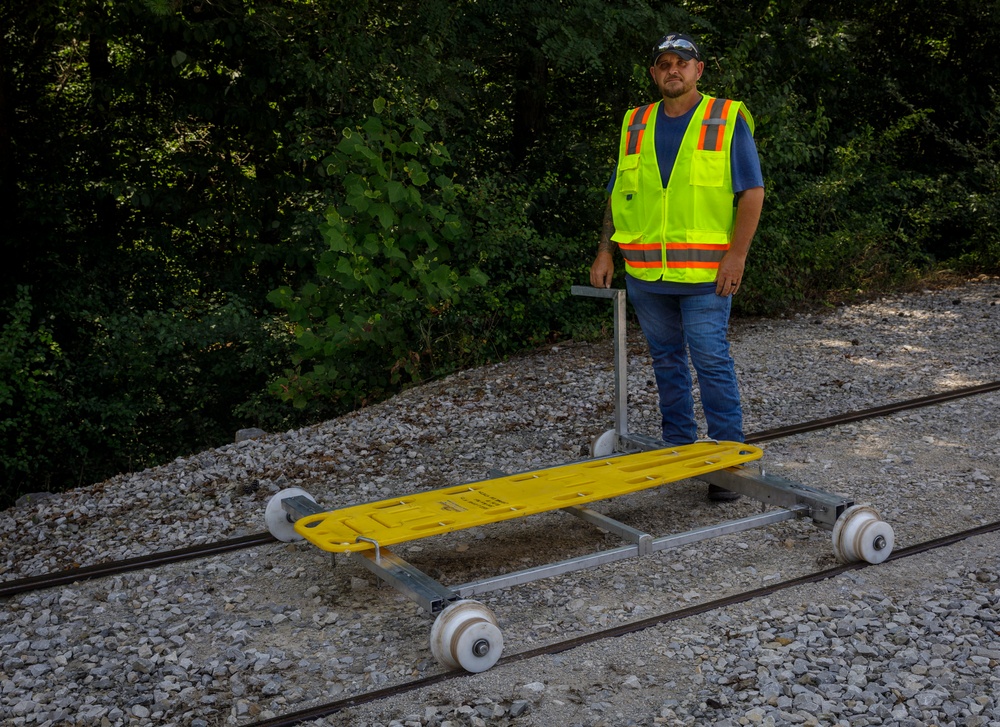 A railroad worker in a high-visibility safety vest stands on a gravel rail bed beside a yellow-topped rail inspection or maintenance cart sitting on two parallel tracks. The cart appears to be a manually operated rail trolley with a metal frame, white flanged wheels, and a yellow plastic platform surface. The setting is an outdoor wooded area with dense green foliage in the background.