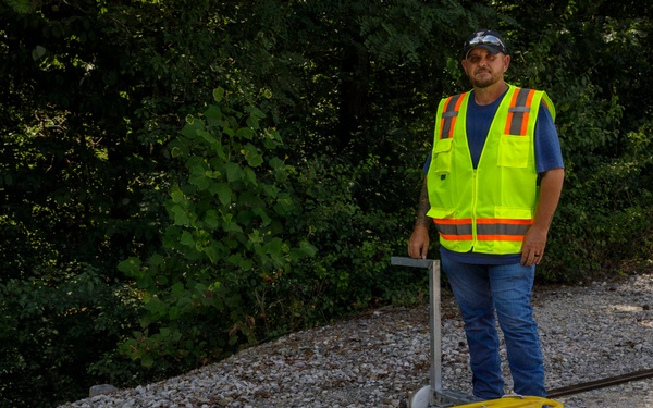 A railroad worker in a high-visibility safety vest stands on a gravel rail bed beside a yellow-topped rail inspection or maintenance cart sitting on two parallel tracks. The cart appears to be a manually operated rail trolley with a metal frame, white flanged wheels, and a yellow plastic platform surface. The setting is an outdoor wooded area with dense green foliage in the background.