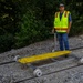 A railroad worker in a high-visibility safety vest stands on a gravel rail bed beside a yellow-topped rail inspection or maintenance cart sitting on two parallel tracks. The cart appears to be a manually operated rail trolley with a metal frame, white flanged wheels, and a yellow plastic platform surface. The setting is an outdoor wooded area with dense green foliage in the background.